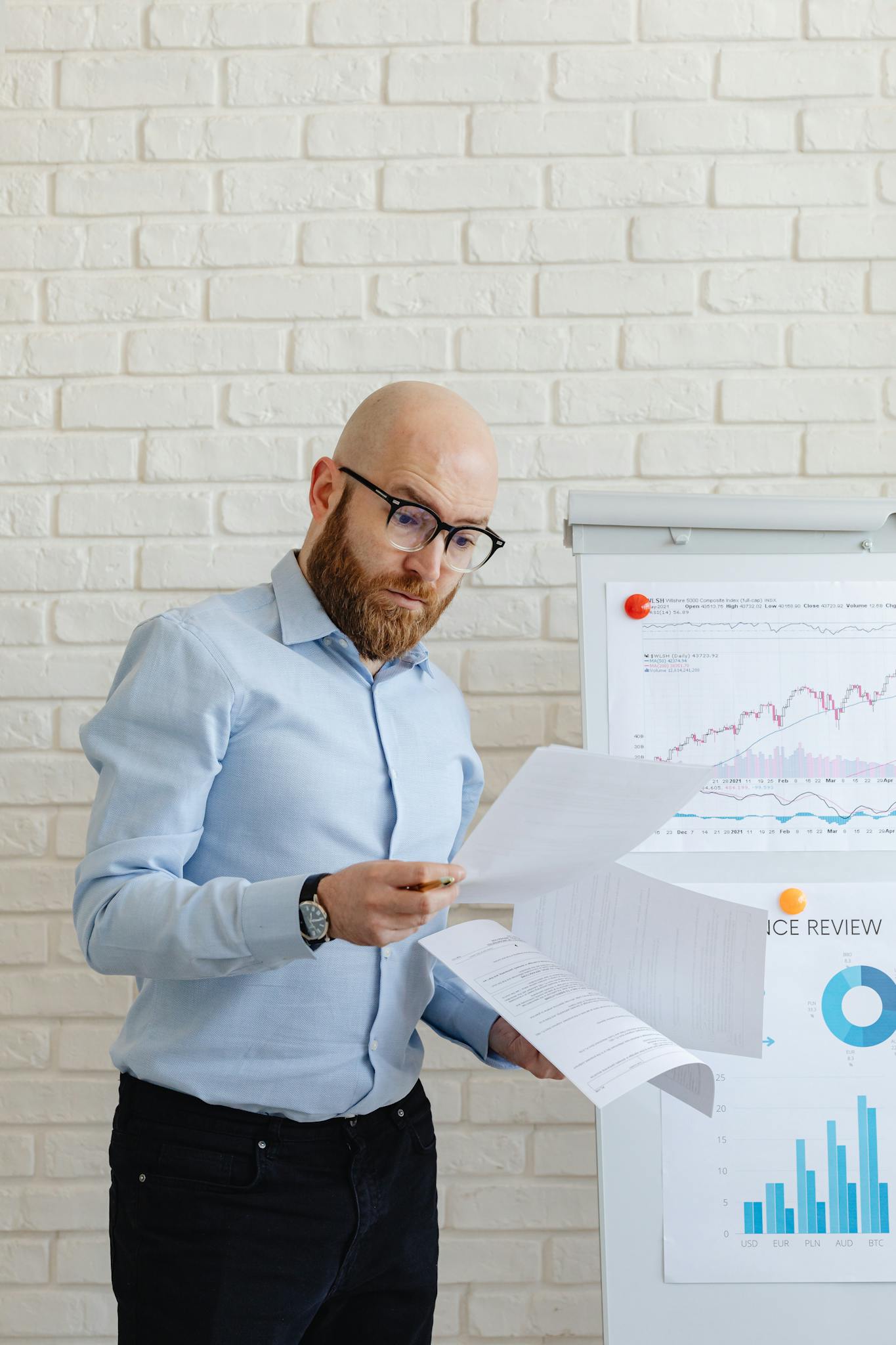 Bald man with eyeglasses reviewing financial papers in front of charts indoors.
